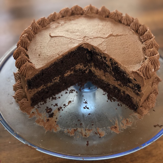 Side view of a round chocolate cake with chocolate buttercream icing with a slice missing to show the layers.