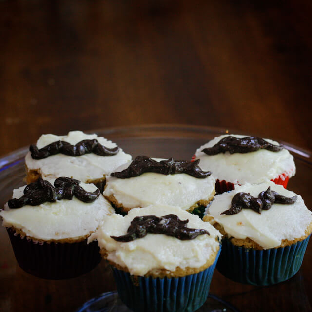 Six vegan Father's Day vanilla cupcakes with buttercream icing decorated with brown mustaches.