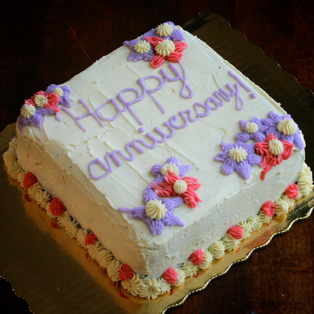 Square cake with white icing, decorated with pink and purple flowers in the corners.