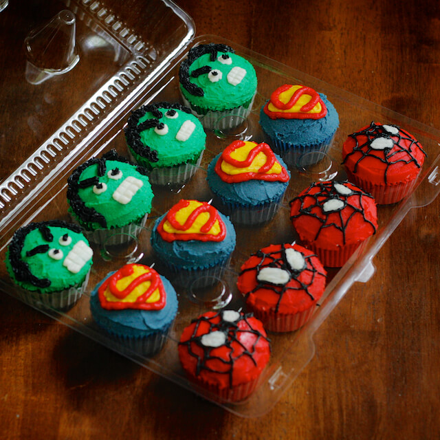 One dozen superhero cupcakes in a clear plastic container. The top row is the Incredible Hulk. The middle row is the Superman Logo. The bottom row is Spiderman.