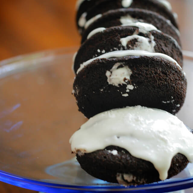 Six chocolate donuts with white cream inside and white icing on a round cake plate.