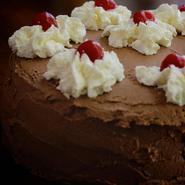 Close-up of a round chocolate cake with chocolate buttercream icing, decorated with whipped cream flowers and cherries.