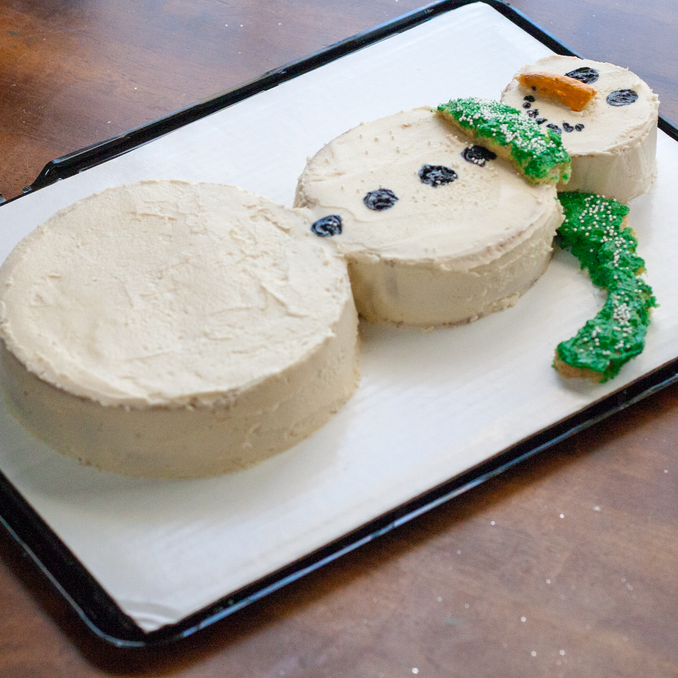 A white snowman cake decorated with black eyes, an orange carrot nose, four black buttons, and a green scarf.